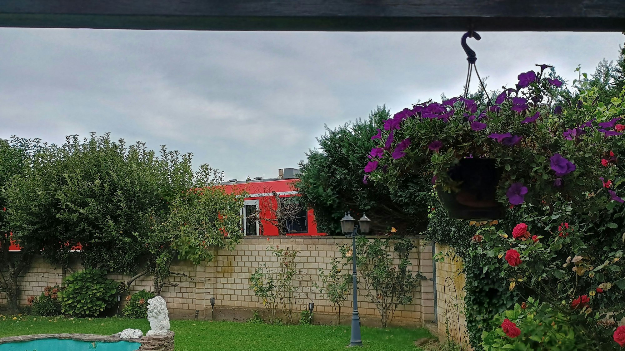 Das Bild zeigt den Garten, hinter der Mauer sieht man den roten Zug zwischen Büschen entlangfahren.