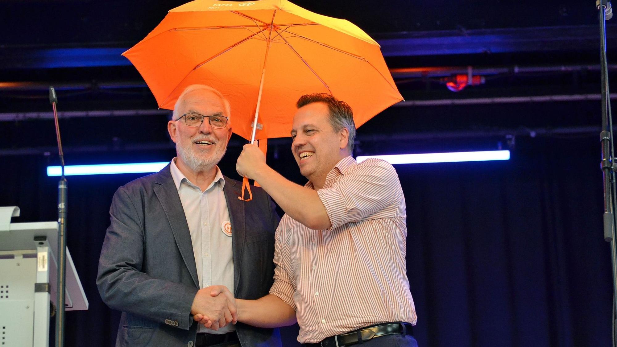 Bürgermeister Sacha Reichelt hält einen orangefarbenen Regenschirm über einen Mann.