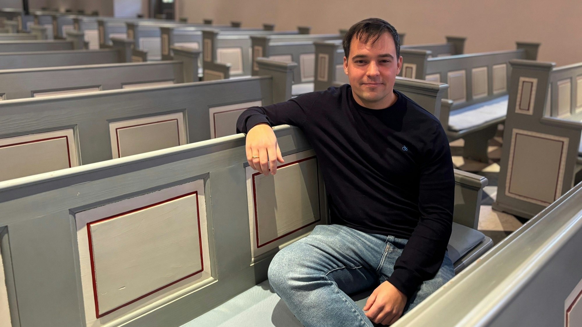Auf dem Bild ist ein Mann in Jeans und Pullover zu sehen, der auf einer Kirchenbank sitzt.