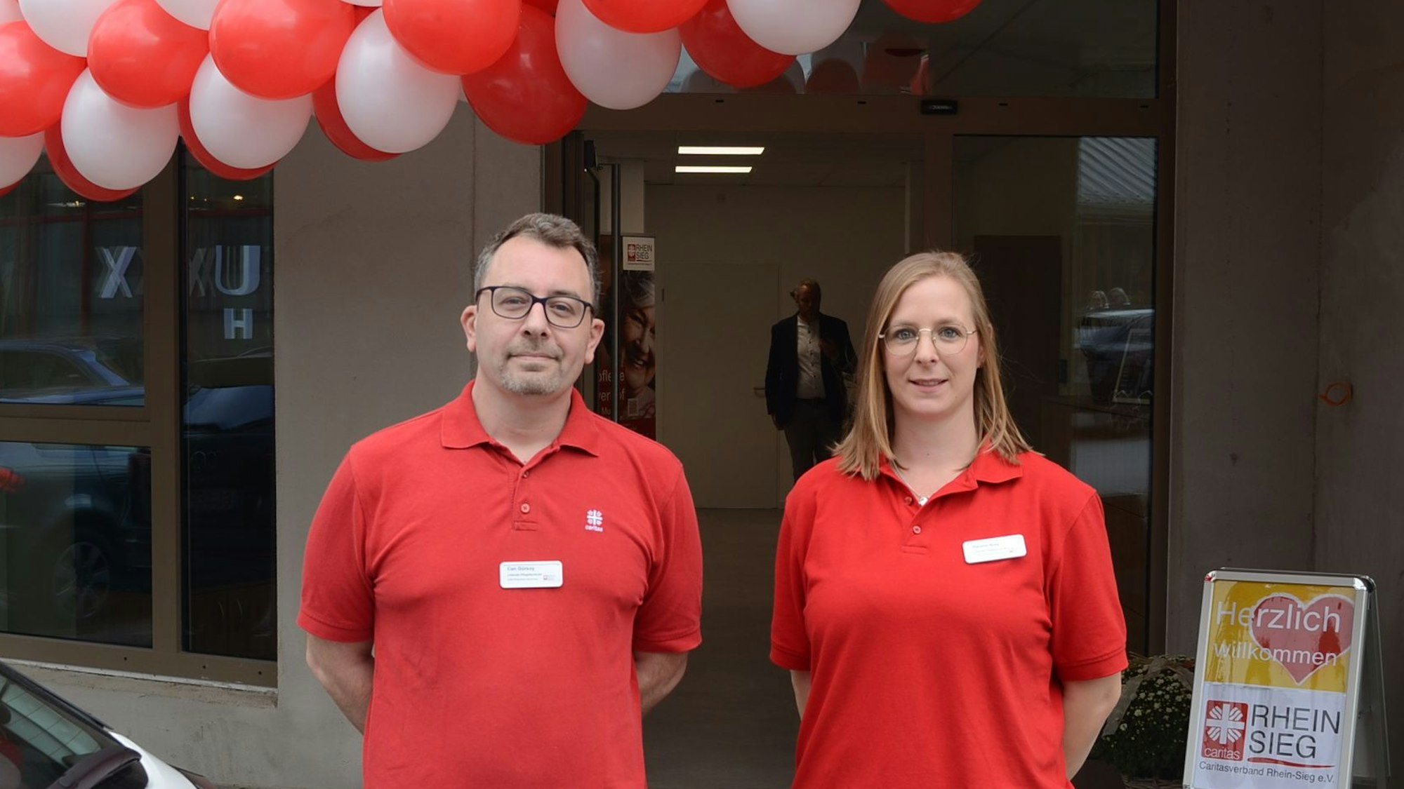 Ein Mann und eine Frau in roten Polohemden; über ihnen hängen rote und weiße Luftballons.