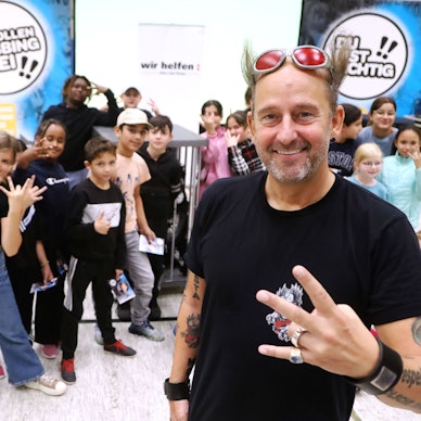 Tom Lehel steht mit 120 Kindern in der Turnhalle der Grundschule Kunterbunt vor Plakaten mit der Aufschrift „wir helfen“ und „Wir wollen mobbingfrei“
