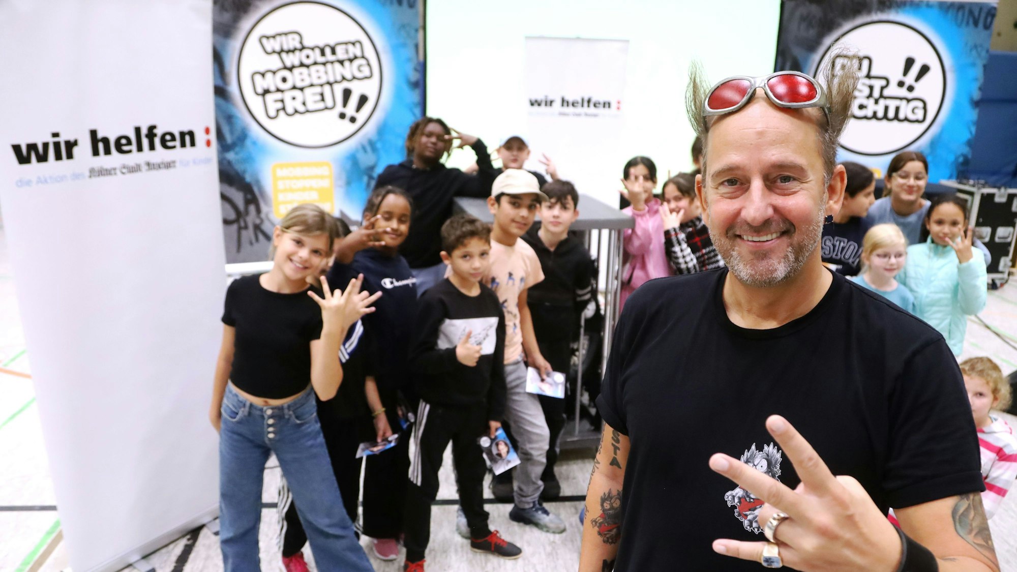 Tom Lehel steht mit 120 Kindern in der Turnhalle der Grundschule Kunterbunt vor Plakaten mit der Aufschrift „wir helfen“ und „Wir wollen mobbingfrei“