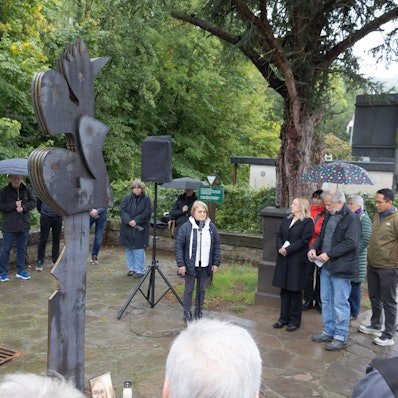 Eine Gruppe von Menschen steht im Halbkreis um eine aus Cortenstahl gefertigte Skulptur herum. Einige haben Regenschirme aufgespannt.