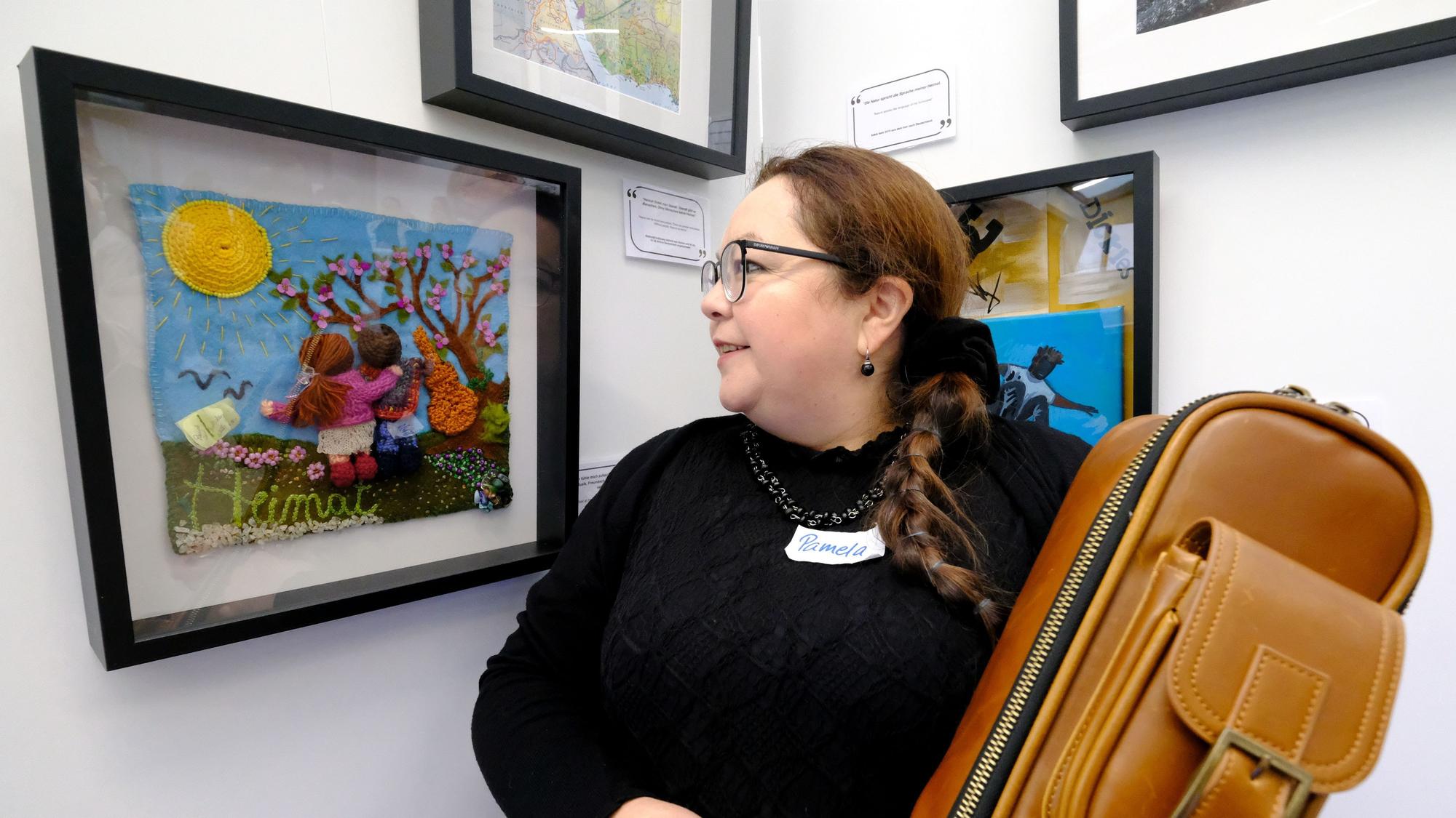 Eine Frau mit Brille und schwarzem Pullover blickt auf ein gerahmtes Kunstwerk, das mit anderen Arbeiten in einer Ausstellung zu sehen ist.