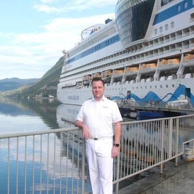 Federico Sass aus Euskirchen ist Chief Purser der Aida-Kreuzfahrtflotte. Hier steht er in seiner weißen Dienstkleidung vor einem Schiff.