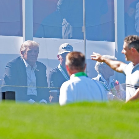 Donald Trump sitzt hinter Plexiglas, während Rory McIlroy ins Publikum grüßt.