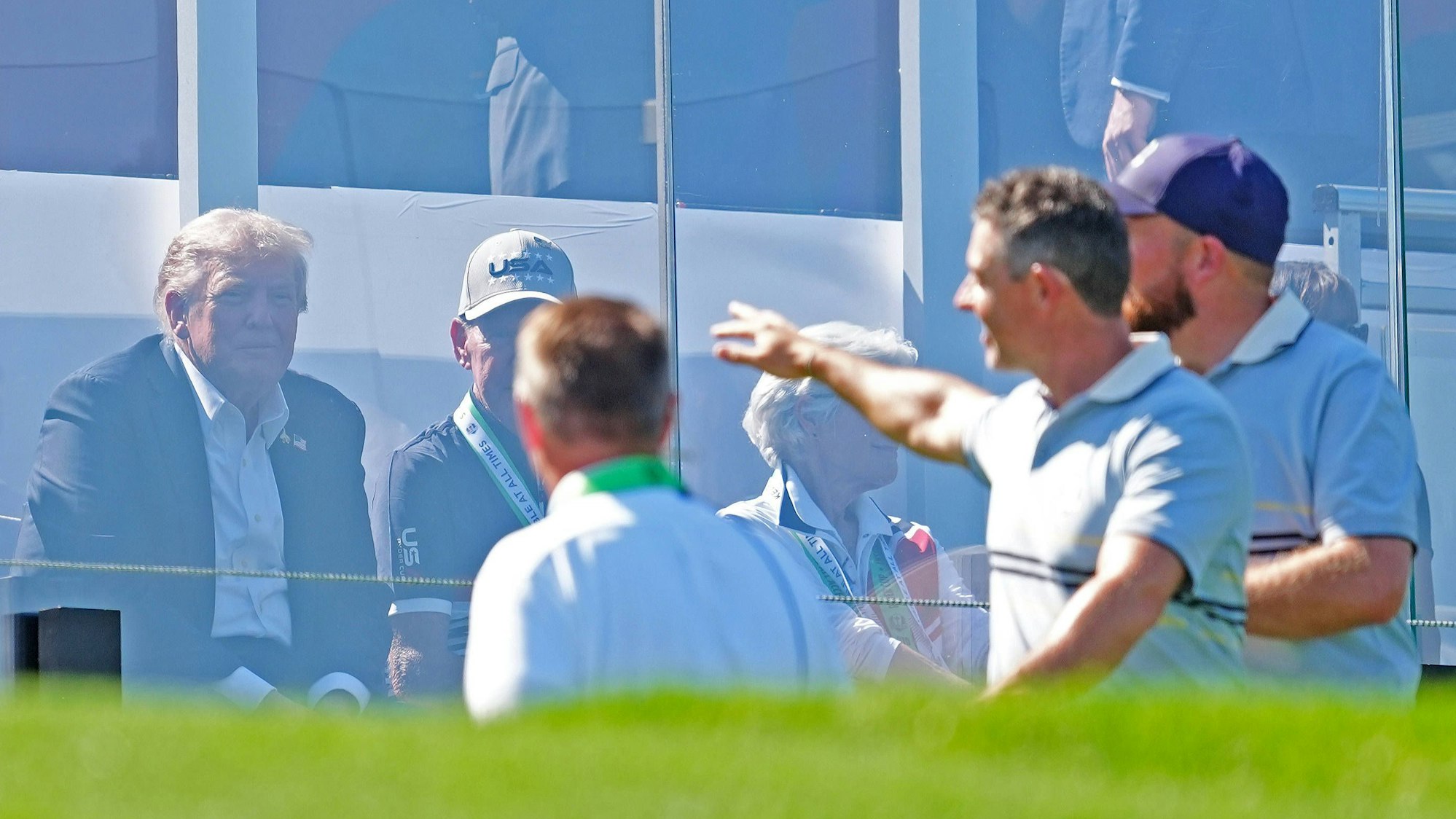 Donald Trump sitzt hinter Plexiglas, während Rory McIlroy ins Publikum grüßt.