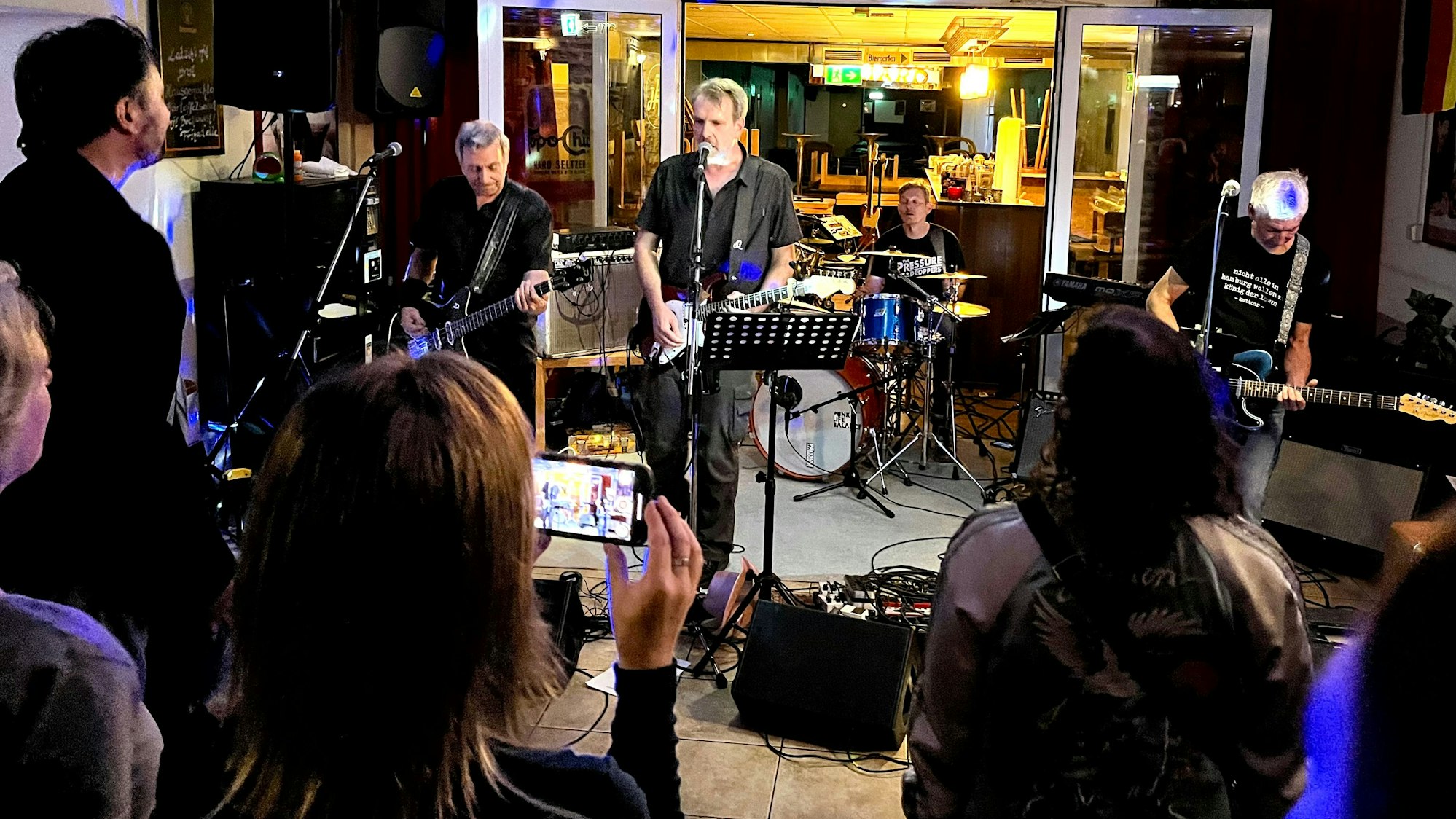 Zu sehen sind eine Band, die in einer Gaststätte spielt, und Zuhörer.