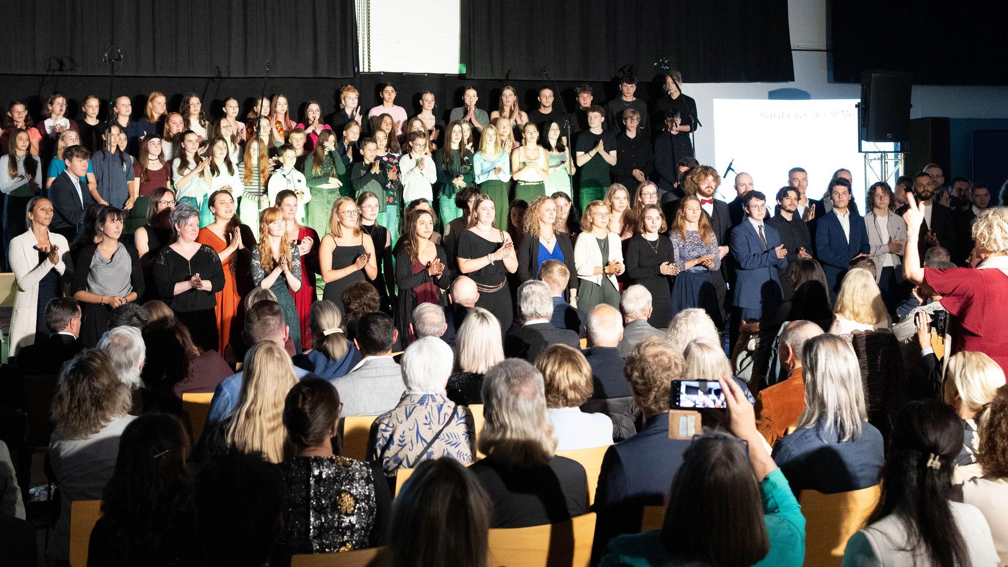 Aktuelle und ehemalige Schüler des St.-Michael-Gymnasiums in Bad Münstereifel stehen auf einer Bühne und singen gemeinsam.
