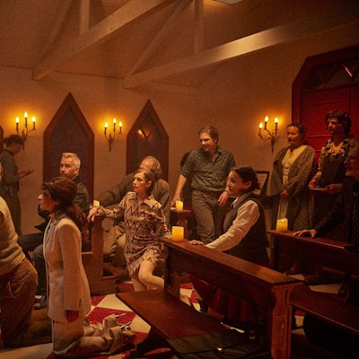 Schauspieler knien in einer Kirche vor einem halbnackten Alten mit Blumenkranz.