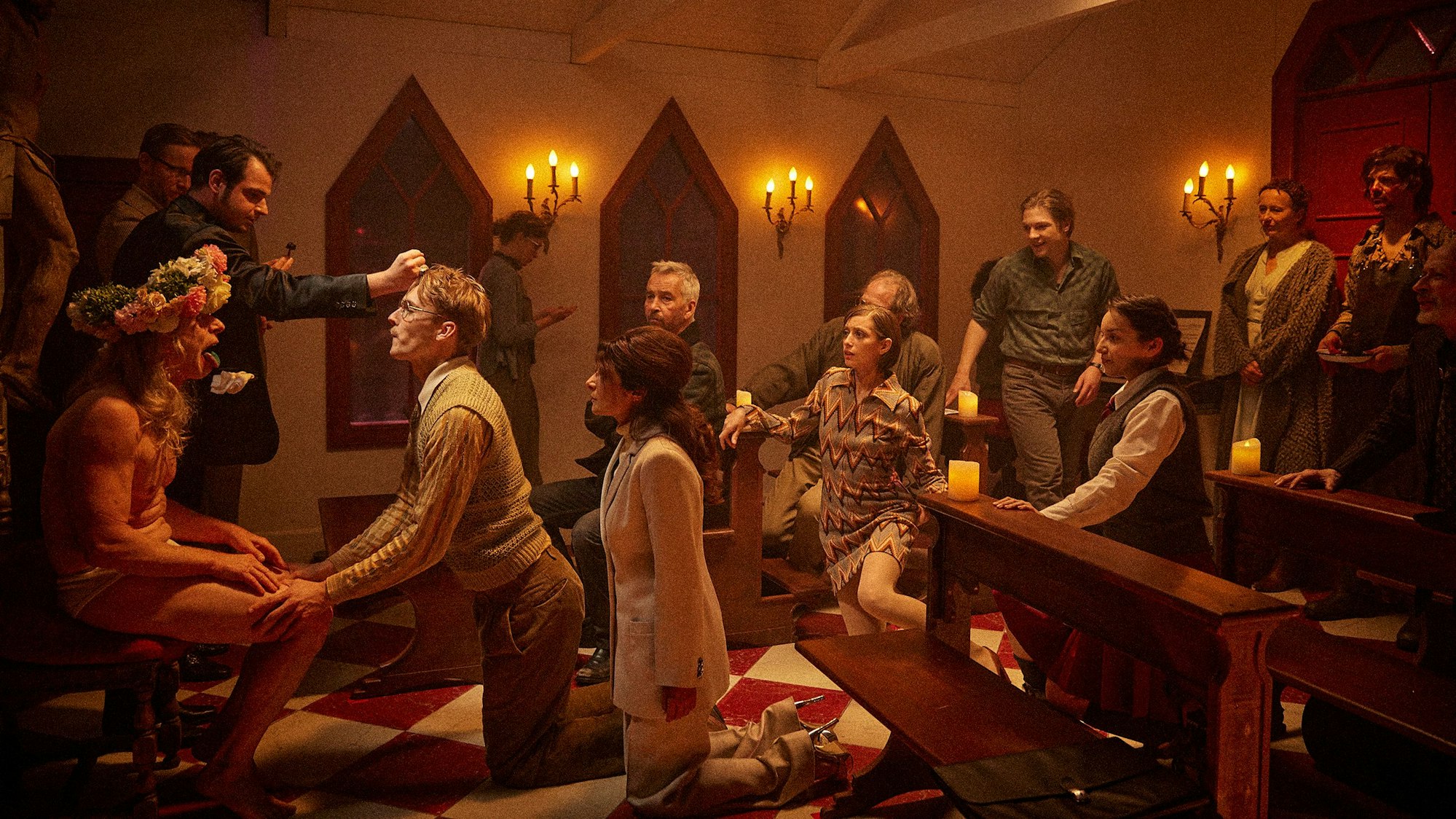 Schauspieler knien in einer Kirche vor einem halbnackten Alten mit Blumenkranz.