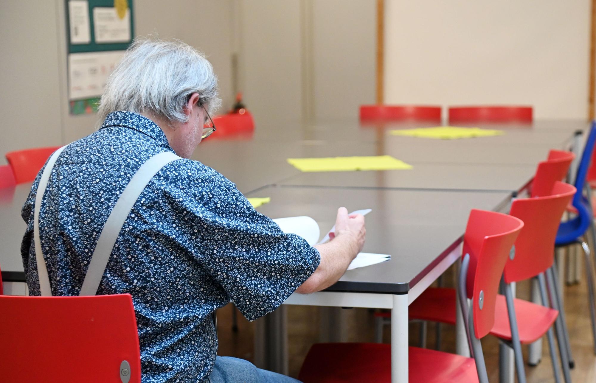 Ein Mann mit grauen Haaren und Brille trägt ein Hemd mit Hosenträgern und schaut auf ein Heft vor ihm, das er umschlägt. Der Mann ist von der Kamera abgewandt, sodass sein Gesicht nicht zu erkennen ist.