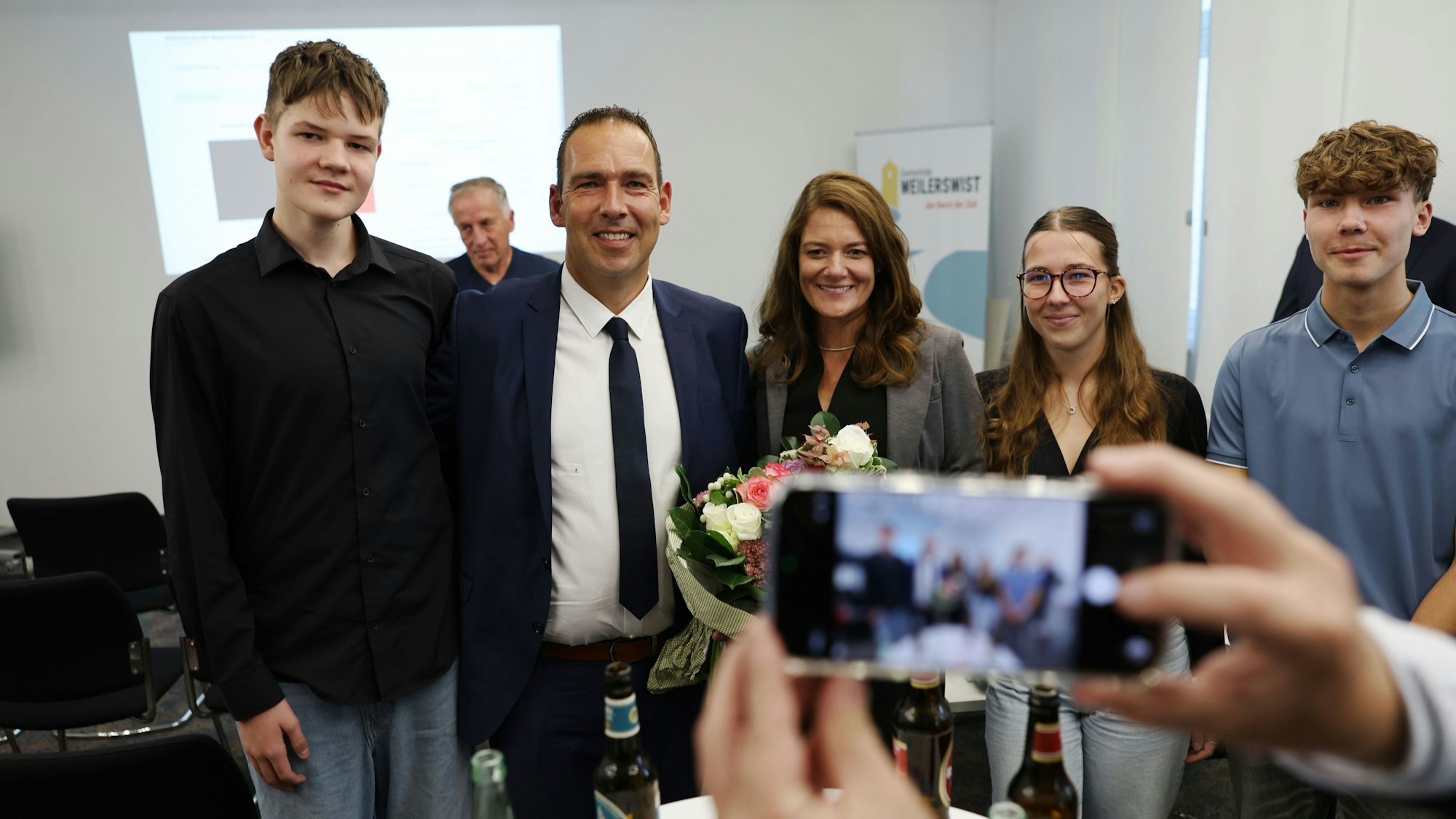 Der künftige Weilerswister Bürgermeister Dino Steuer steht mit seiner Frau Julia, Tochter Lara-Sophie und den beiden Söhnen Ben (l.) und Jan zusammen. Im Vordergrund ist ein Smartphone zu sehen, mit dem ein Foto der Familie gemacht wird