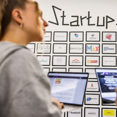 Teilnehmer eines Meetings sprechen im Start-up-Hub «STARTPLATZ» miteinander, während im Hintergrund an einer Wand mit dem Schriftzug «Startups» die Logos junger Unternehmen zu sehen sind.