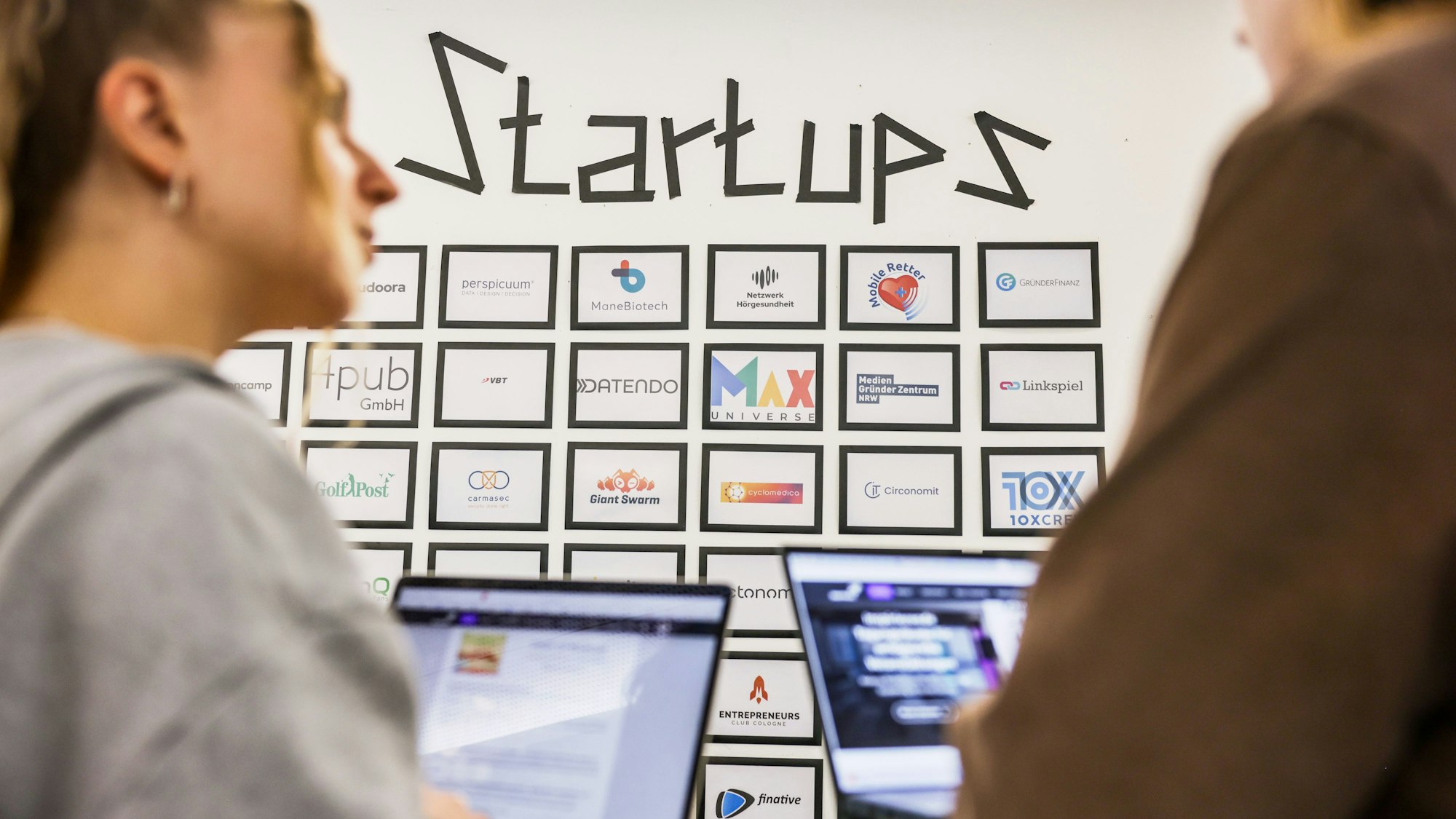 Teilnehmer eines Meetings sprechen im Start-up-Hub «STARTPLATZ» miteinander, während im Hintergrund an einer Wand mit dem Schriftzug «Startups» die Logos junger Unternehmen zu sehen sind.