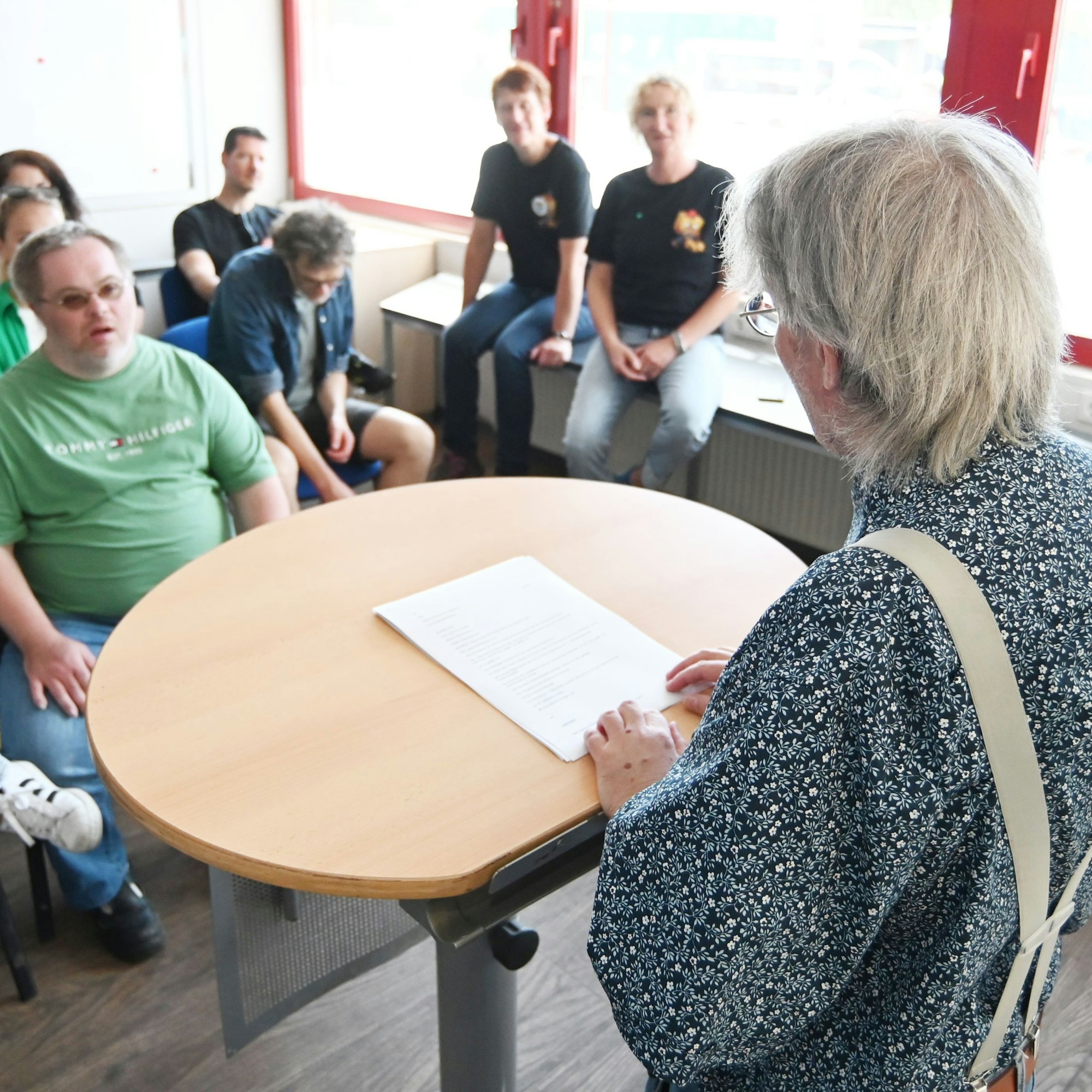 Die Aufnahme wurde so gemacht, dass Andreas K. schräg zur Kamera steht und nicht zu erkennen ist. Im Gegenüber sitzen mehrere Menschen, die seiner Lesung zuhören. Sie sind unscharf gestellt.