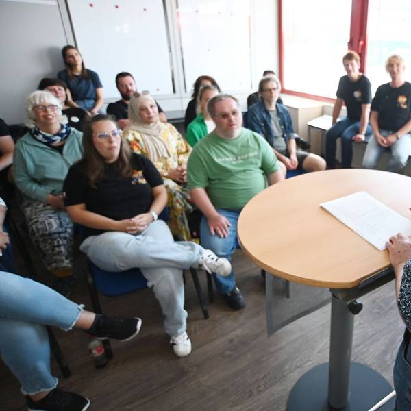 Herr K. trägt Hemd und Hosenträger und liest von einem Blattpapier ab, das auf einem Rednerpult liegt. Aufgrund der seitlichen Perspektive, ist er im Gesicht nicht zu erkennen. Vor ihm sitzen Zuhörer und Zuhörerinnen, die unscharf gestellt wurden.