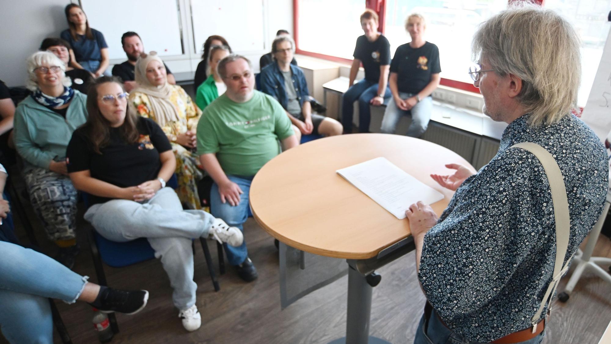 Herr K. trägt Hemd und Hosenträger und liest von einem Blattpapier ab, das auf einem Rednerpult liegt. Aufgrund der seitlichen Perspektive, ist er im Gesicht nicht zu erkennen. Vor ihm sitzen Zuhörer und Zuhörerinnen, die unscharf gestellt wurden.