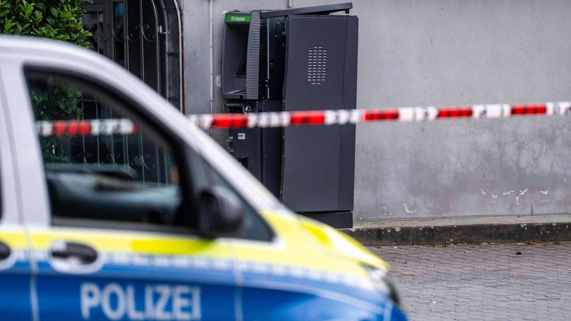 Das Bild zeigt einen Streifenwagen, der von einem Flatterband steht. Im Hintergrund ist der zerstörte Geldautomat zu sehen.