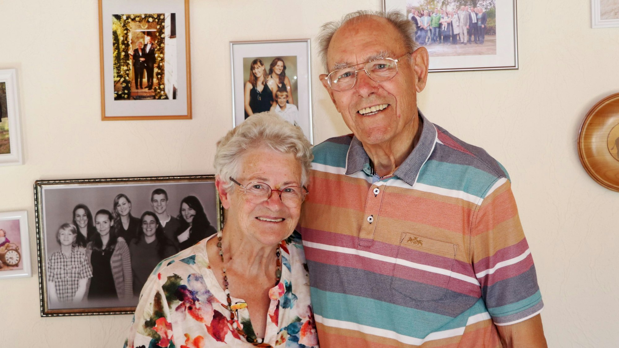 Das Bild zeigt das Ehepaar, sie hat eine bunt geblümte Bluse an, er hat ein buntes gestreiftes Hemd. An der Wand hinter ihnen hängen Fotos.