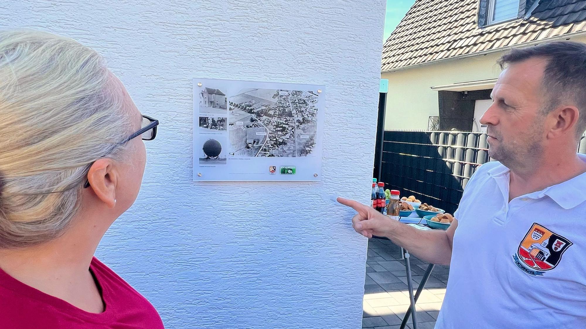 Ein Mann und eine Frau betrachten eine Fototafel, die an einer Hauswand angebracht ist.