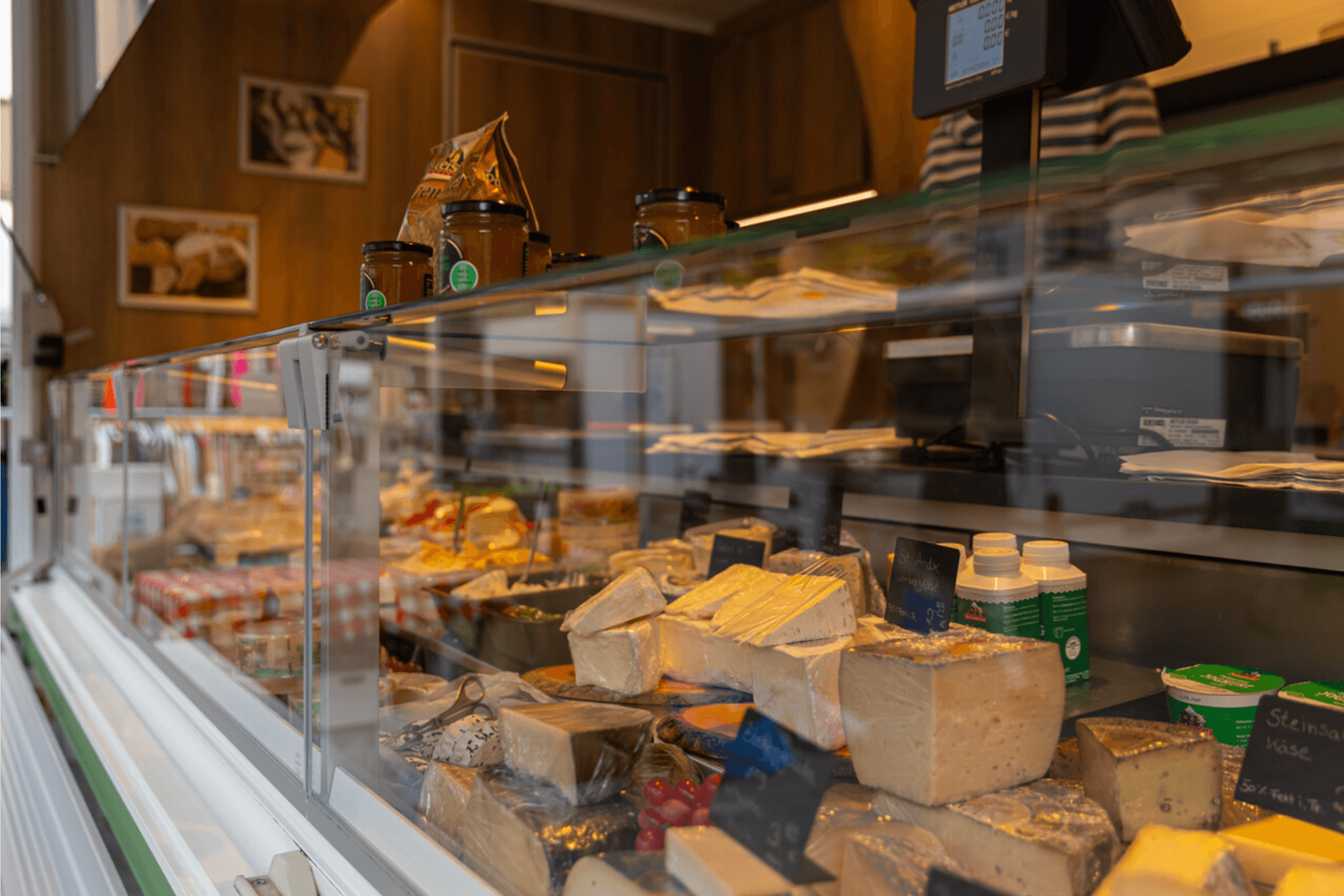 Das Bild zeigt eine Käsetheke auf einem Marktstand, gefüllt mit einer großen Auswahl an Käsesorten – von mild bis kräftig. Hinter der Glasscheibe sind verschiedene Laibe, Stücke und Spezialitäten kunstvoll angerichtet, die zum Probieren und Kaufen einladen.