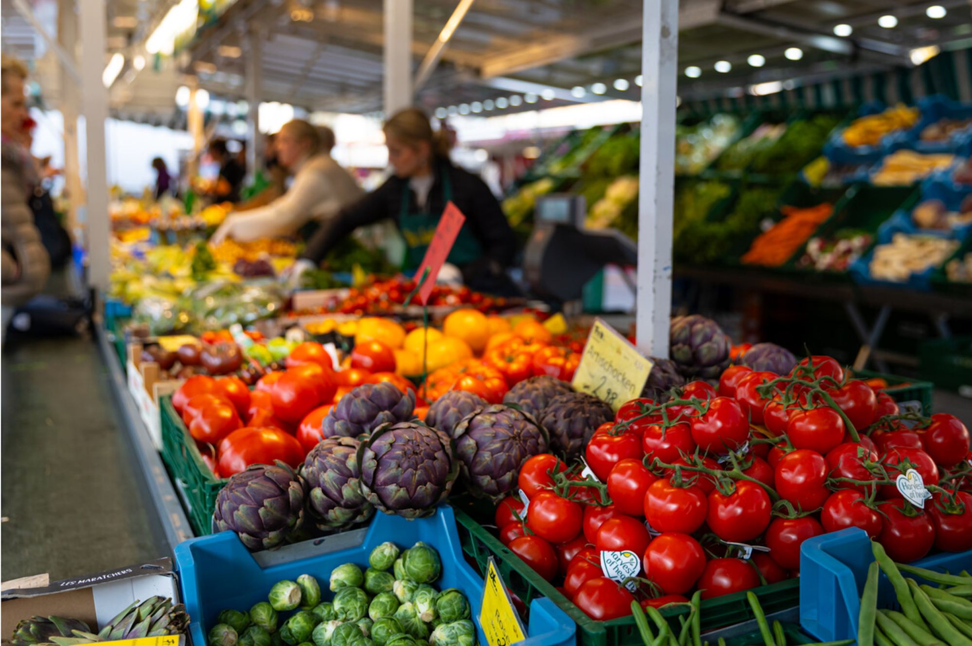 Das Bild zeigt einen bunten Marktstand mit frischem Gemüse wie Tomaten, Artischocken, Rosenkohl und Paprika. Im Hintergrund bedienen Händlerinnen die Kundschaft.