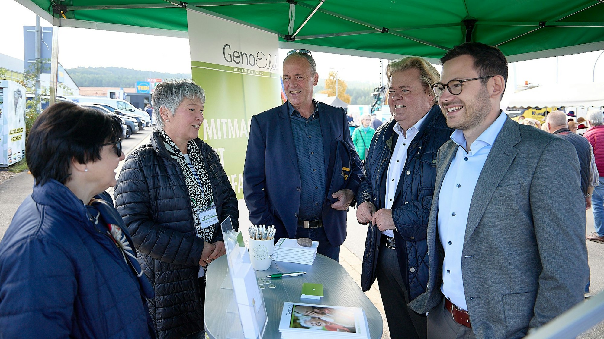 Drei Männer und zwei Frauen stehen an einem Infostand, im Hintergrund ist der Schriftzug Geno Eifel zu sehen.