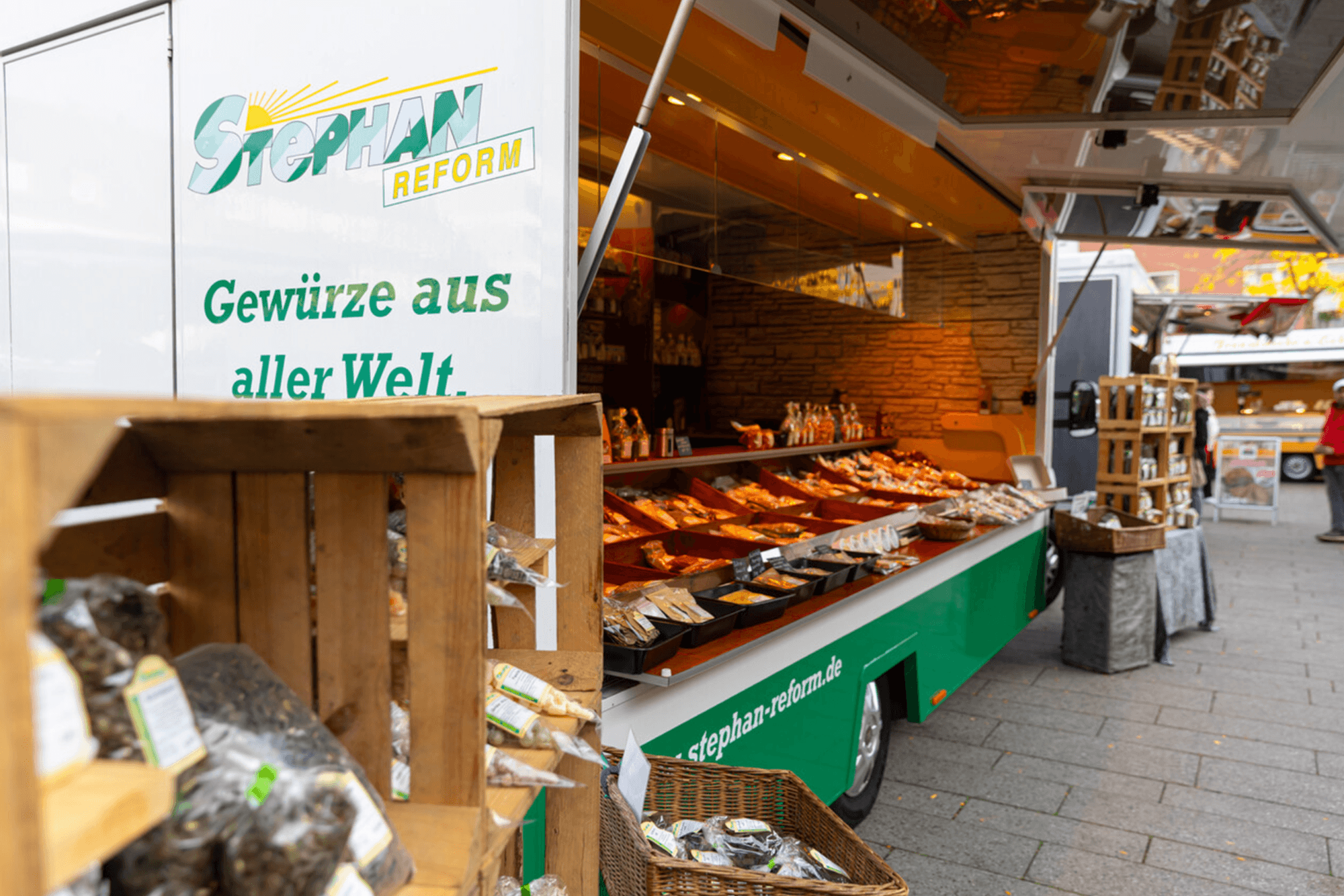 Auf dem Bild ist ein Marktstand zu sehen, der mit dem Slogan „Gewürze aus aller Welt“ wirbt. In Holzkisten und Auslagen werden zahlreiche verpackte Gewürze präsentiert, die zum Stöbern und Kaufen einladen.