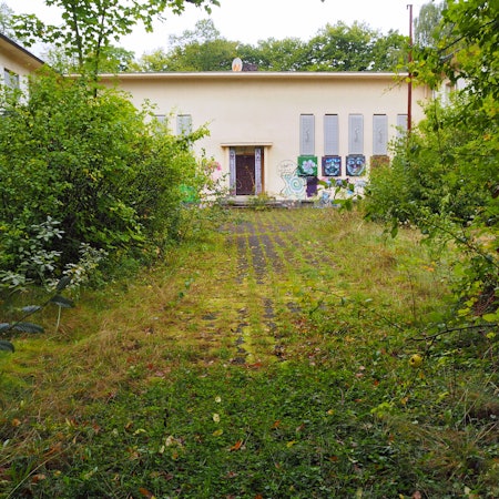 Ein beiges Gebäude mit Flachdach steht hinter Büschen und einer zugewucherten Asphaltfläche.