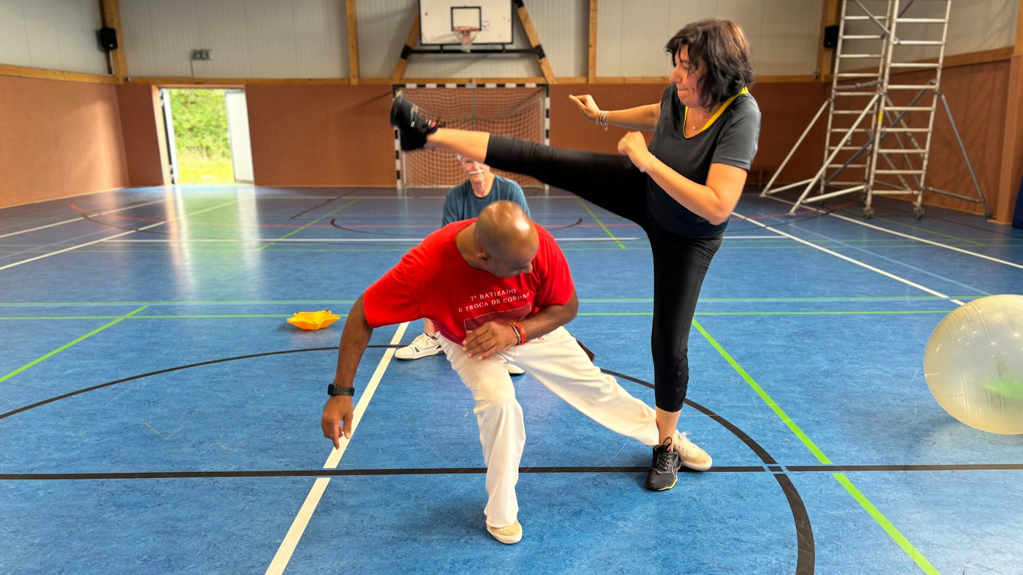 Eine Frau tritt über einen Mann, der sich unter ihren Tritt duckt und gegen ihr Standbein tritt.