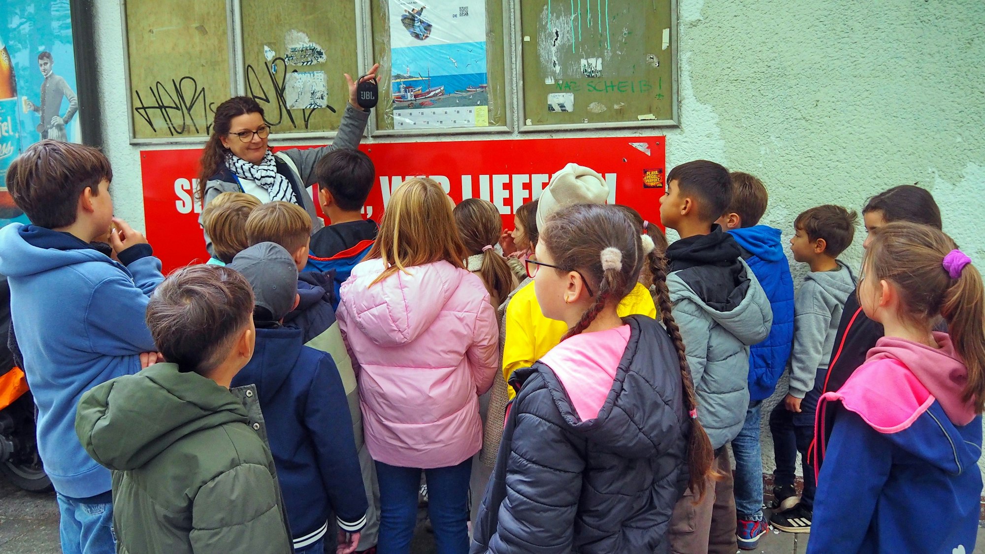 Eine Gruppe Schüler steht vor einem Plakat. Ihre Lehrerin zeigt darauf.