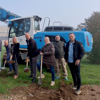 Einige Frauen und Männer stehen vor einem blauen Bagger. Sie haben Schaufeln und Spaten in der Hand, um den symbolischen ersten Spatenstich für die Erweiterung der Grundschule in Blankenheim auszuführen.