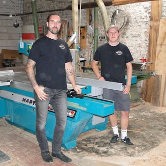 Zwei junge Männer in schwarzen T-Shirts stehen an einer großen Säge in einer Werkstatt.