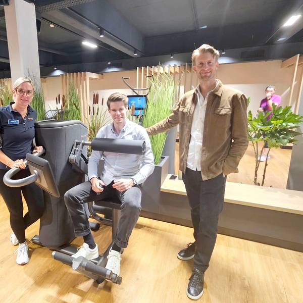 Henning Förster steht neben einem Fitnessgerät, in dem sein Bruder Steffen sitzt. Daneben steht Tanja Hölz.
