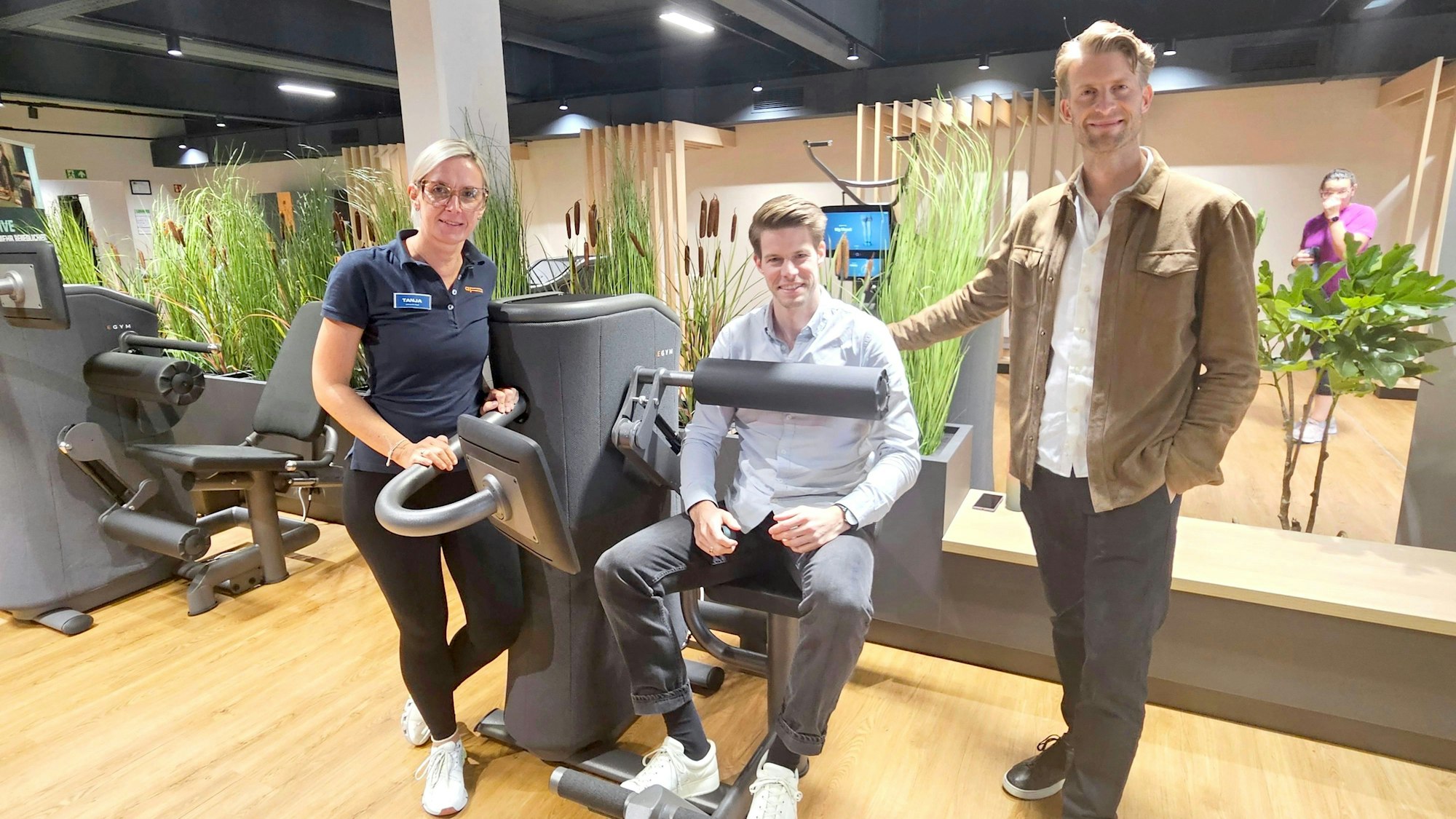 Henning Förster steht neben einem Fitnessgerät, in dem sein Bruder Steffen sitzt. Daneben steht Tanja Hölz.