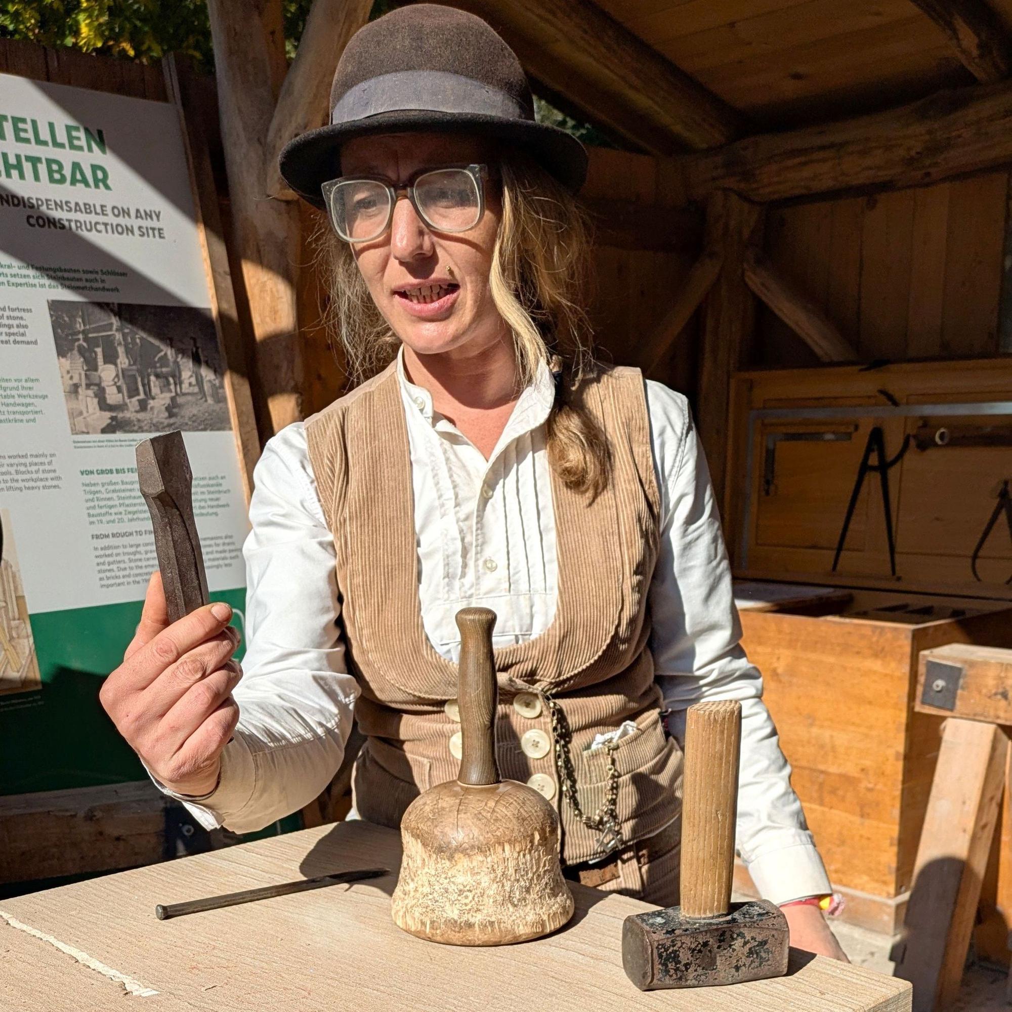 Die Steinmetzmeisterin trägt einen Hut und Arbeitskleidung. Sie hält ein Werkzeug in die Höhe.