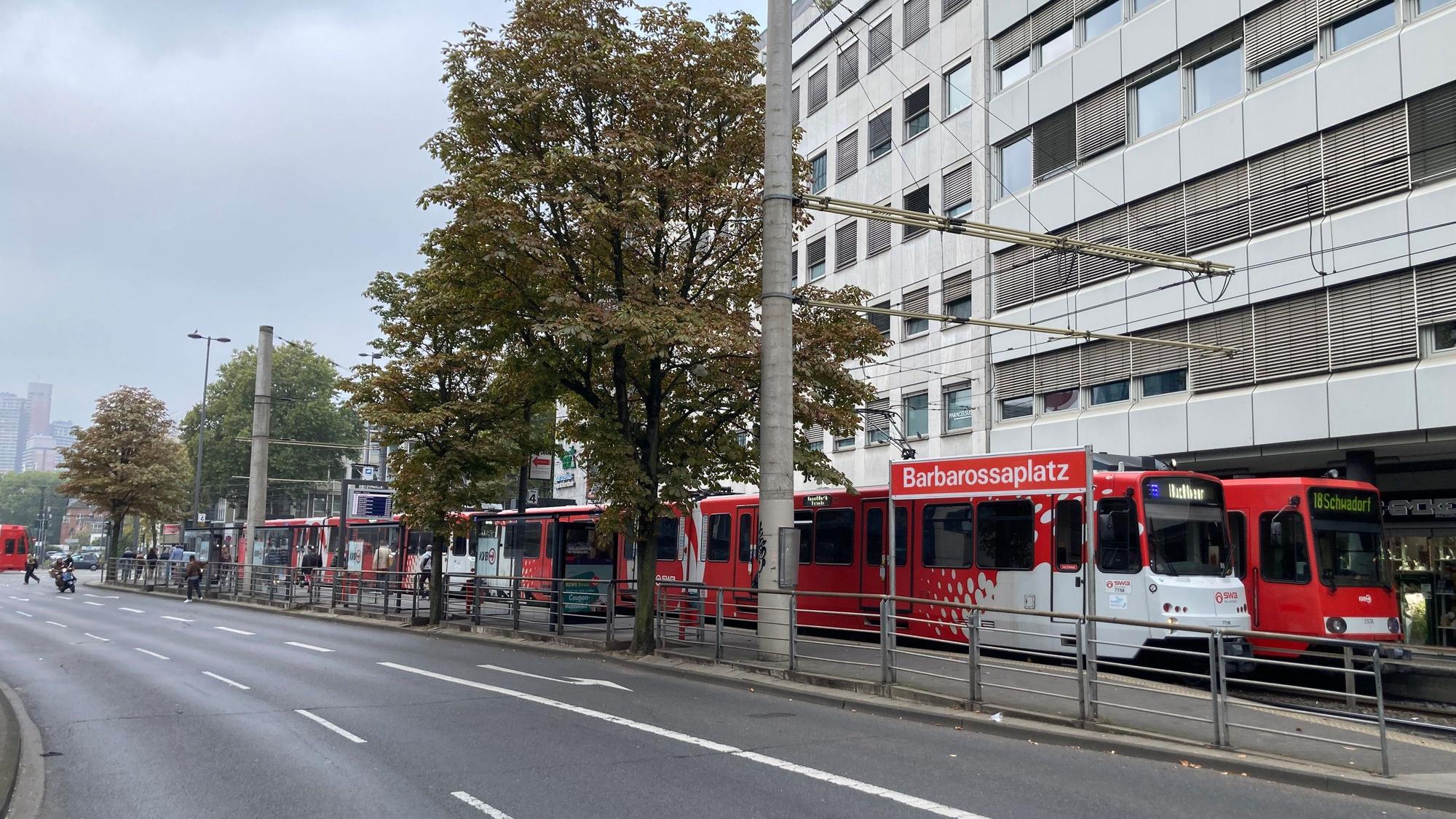 An einer mehrspurigen Straße ist die Haltestelle Barbarossaplatz, an der gerade auch zwei KVB-Züge stehen.