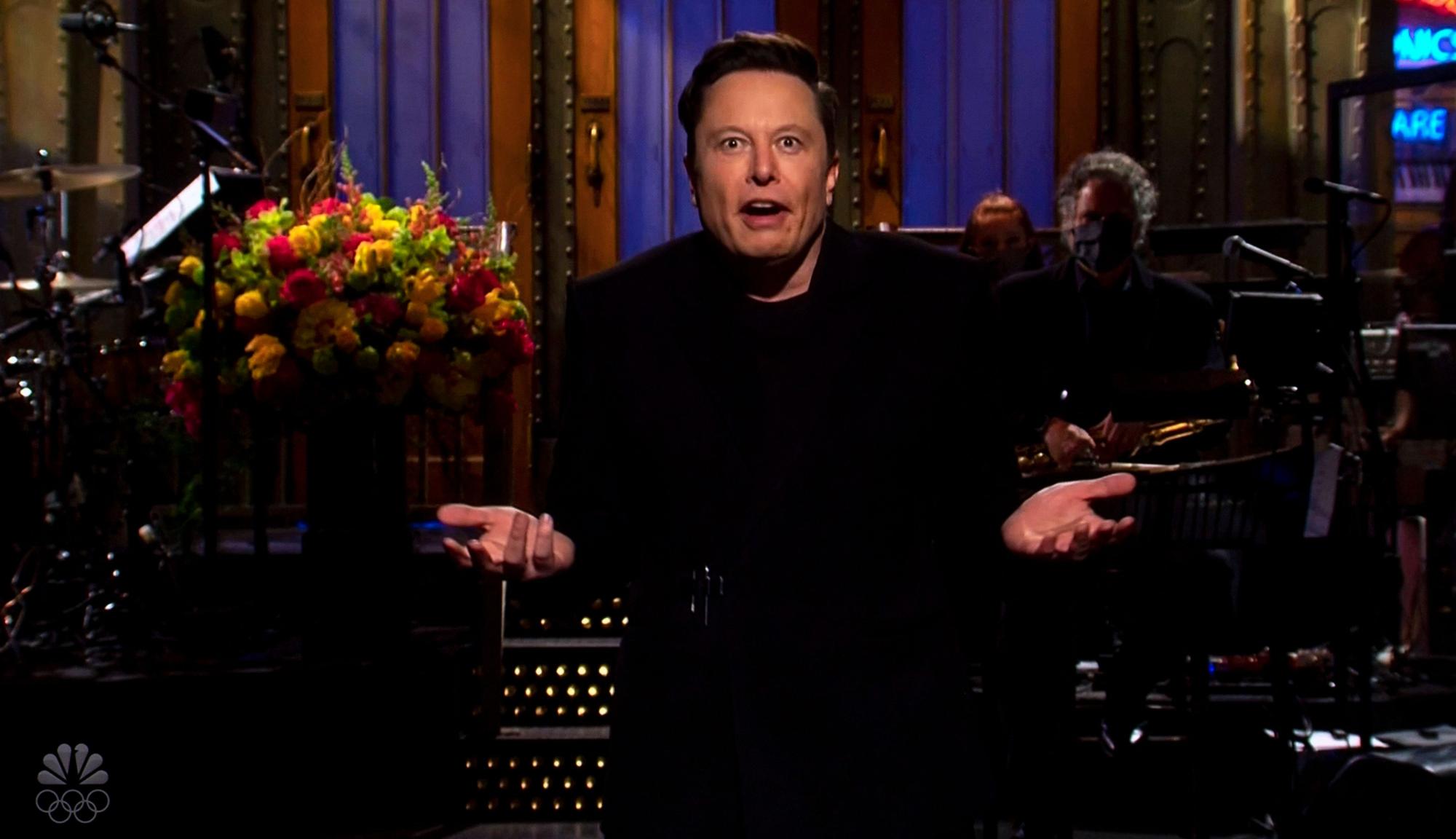 Elon Musk auf einer Bühne, gestikulierend, mit Blumen und Band im Hintergrund.