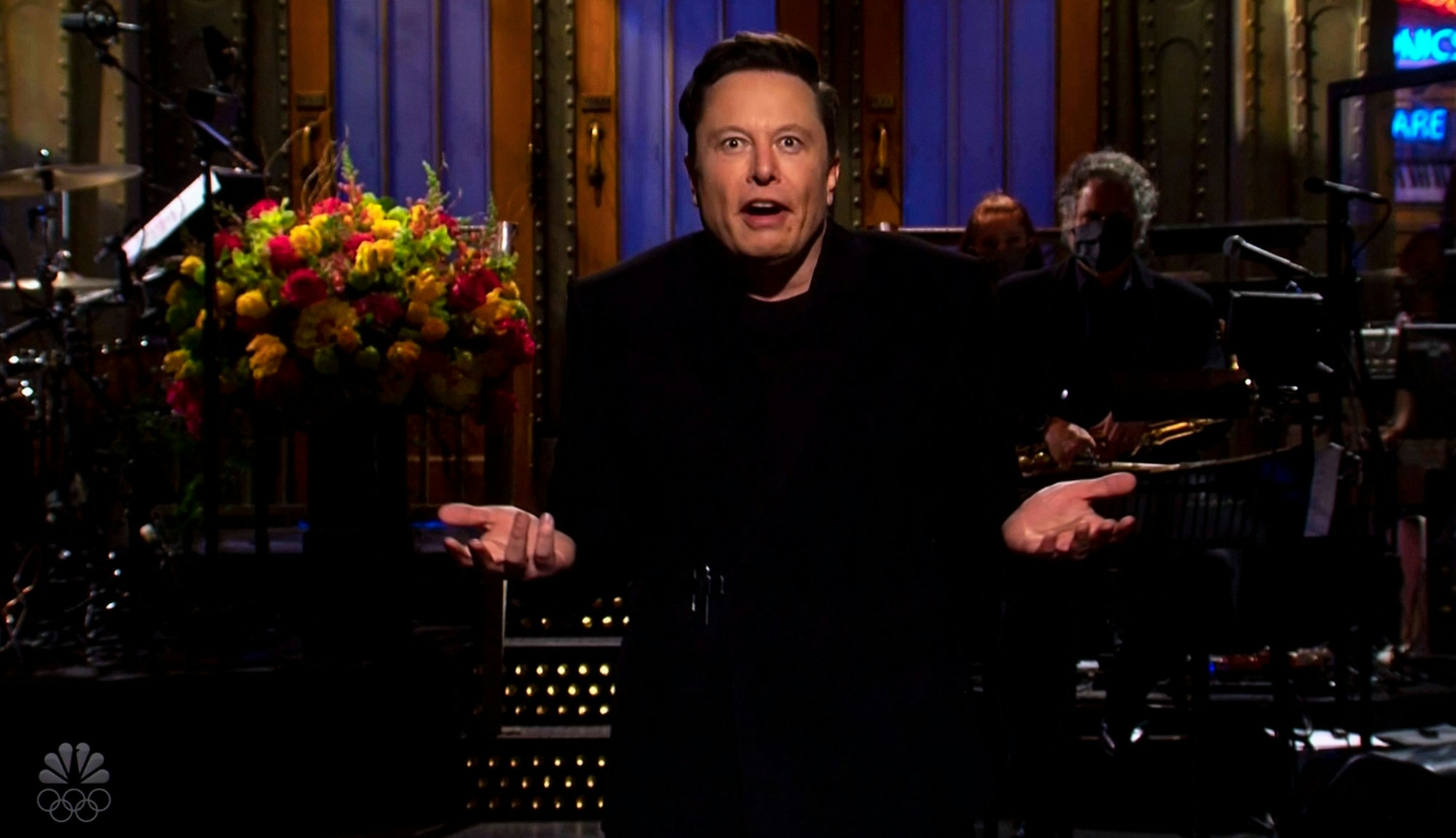 Elon Musk auf einer Bühne, gestikulierend, mit Blumen und Band im Hintergrund.