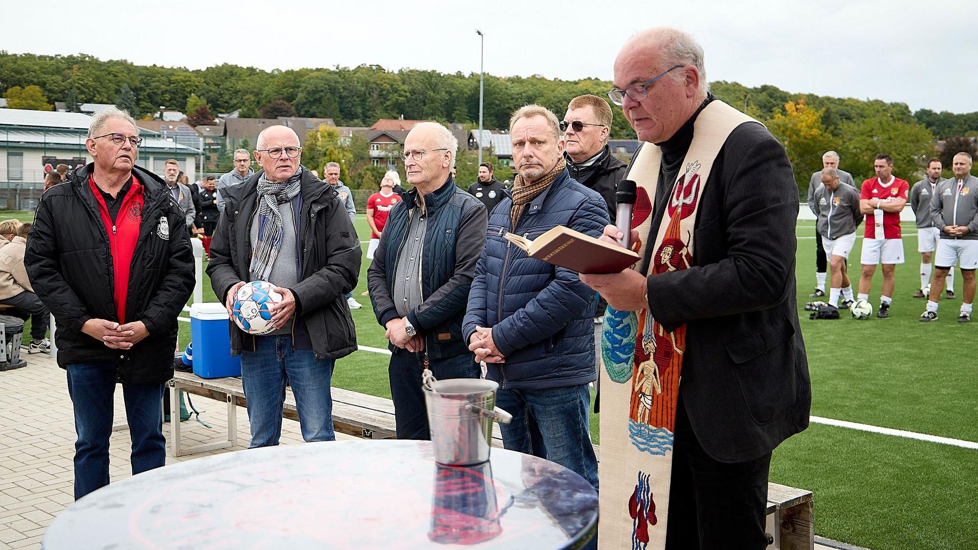 Pastor Erik Pühringer steht mit mehreren Männern an einem Tisch am Rande eines Kunstrasenplatzes.