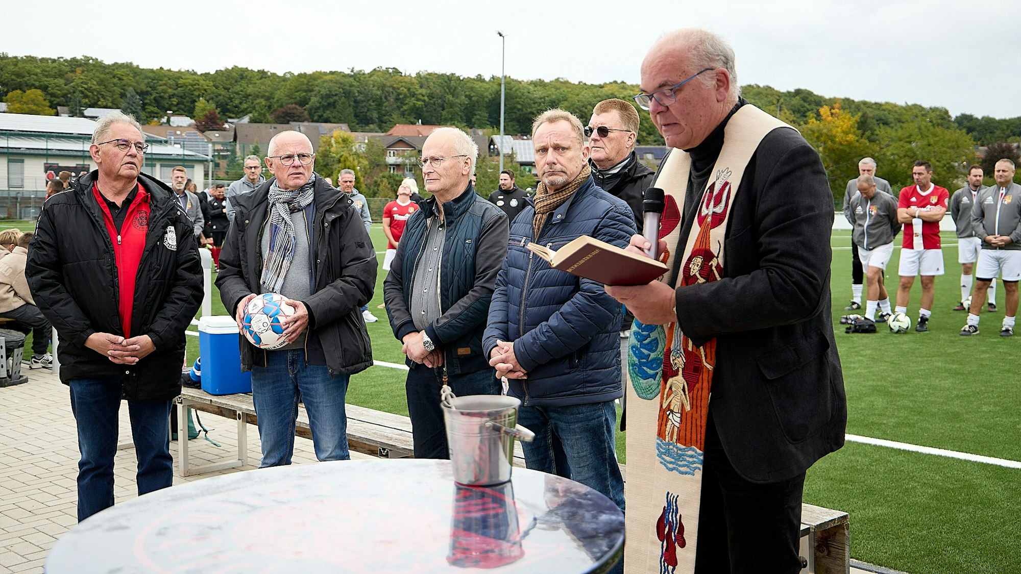 Pastor Erik Pühringer steht mit mehreren Männern an einem Tisch am Rande eines Kunstrasenplatzes.