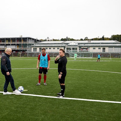 Bürgermeister Dr. Hans-Peter Schick (l.), der dunkle Freizeitkleidung und weiße Sneaker trägt, mit einem Fußballer und dem Schiedsrichter am Mittelkreis des Kunstrasenplatzes in Mechernich.