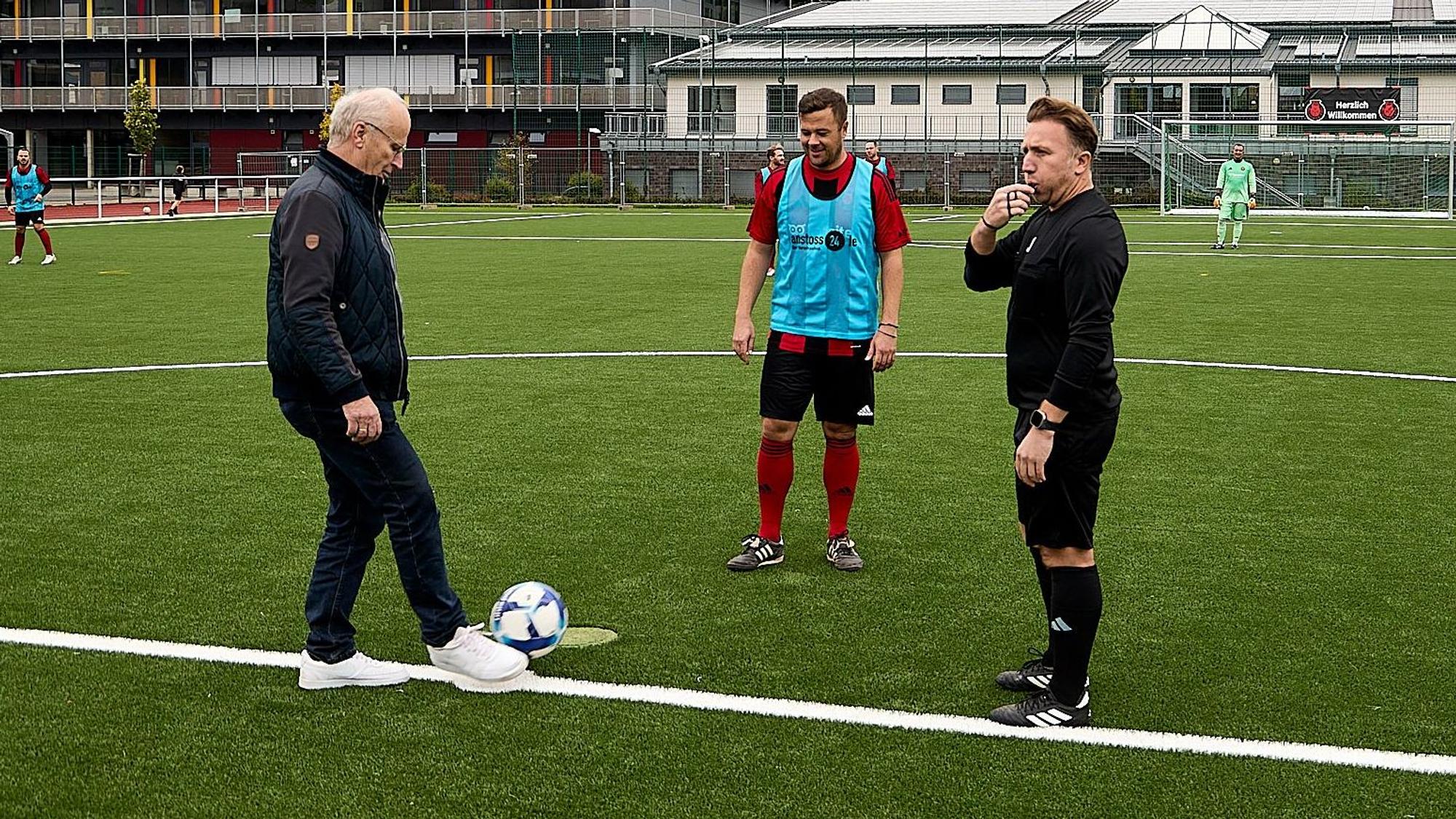 Bürgermeister Dr. Hans-Peter Schick (l.), der dunkle Freizeitkleidung und weiße Sneaker trägt, mit einem Fußballer und dem Schiedsrichter am Mittelkreis des Kunstrasenplatzes in Mechernich.