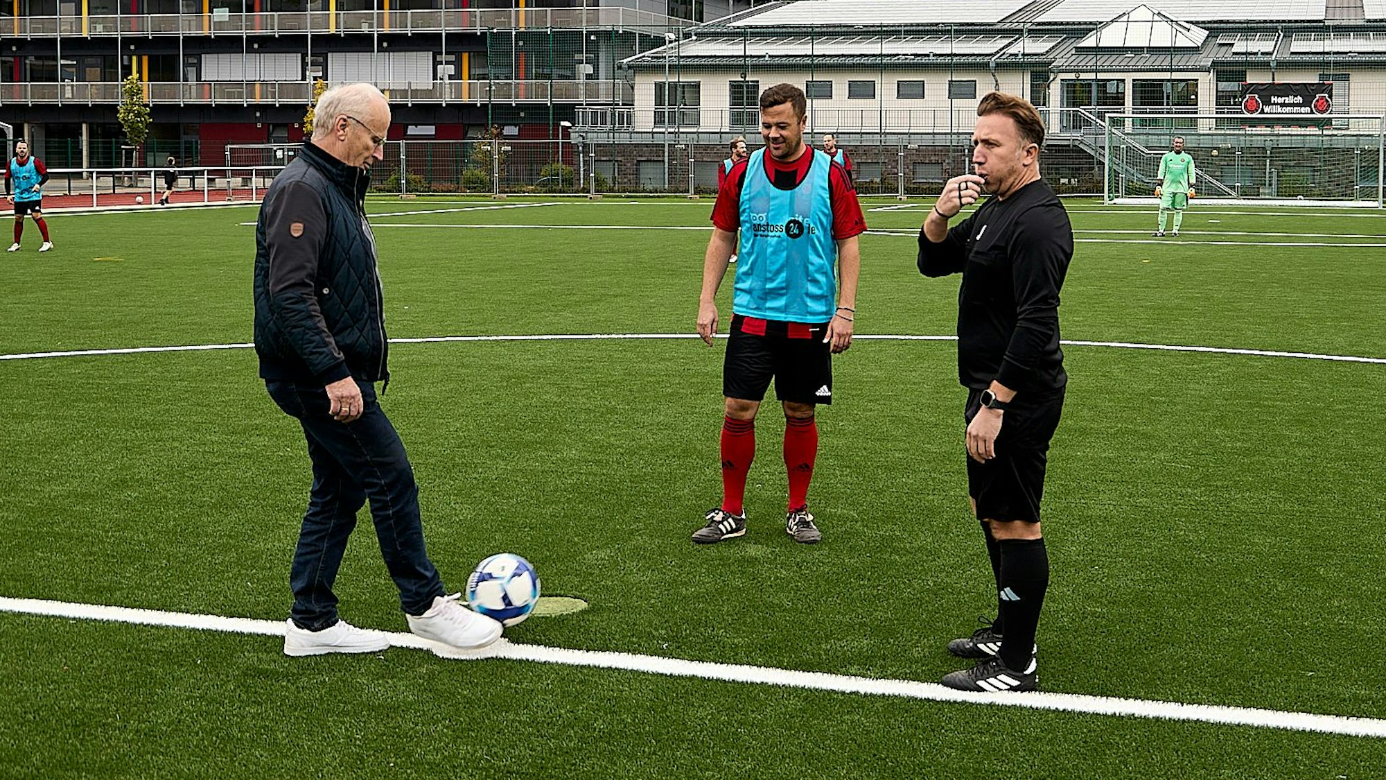 Bürgermeister Dr. Hans-Peter Schick (l.), der dunkle Freizeitkleidung und weiße Sneaker trägt, mit einem Fußballer und dem Schiedsrichter am Mittelkreis des Kunstrasenplatzes in Mechernich.