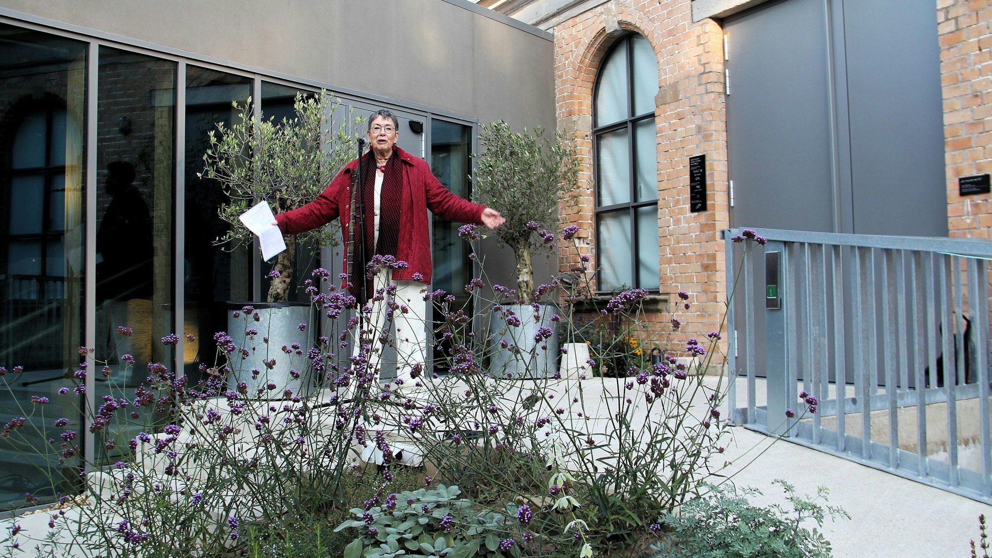 Eine Frau in roter Jacke steht mit ausgebreiteten Armen vor einer Stahl-Glas-Front.