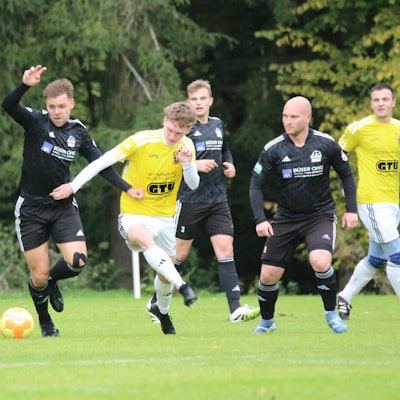 Zwei Fußballer kämpfen um den Ball, mehrere Mitspieler beobachten das Duell.