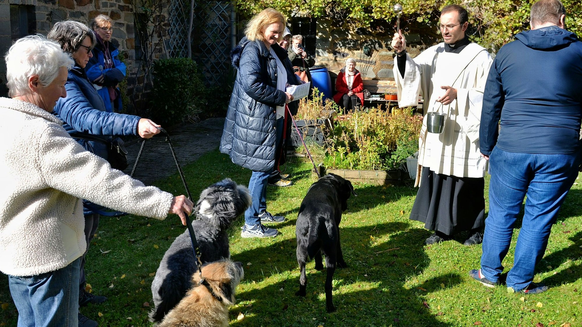 Diakon Francesco Tabacco, der ein liturgisches Gewand trägt, besprengt mehrere Hunde, die von ihren Frauchen an der Leine gehalten werden, mit Weihwasser.
