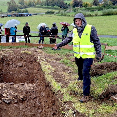 Eine Frau, die eine gelbe Warnweste über ihrer Kleidung trägt, deutet auf eine offene Ausgrabungsstelle auf einer Wiese, um die sich mehrere Besucher mit Regenschirmen versammelt haben.