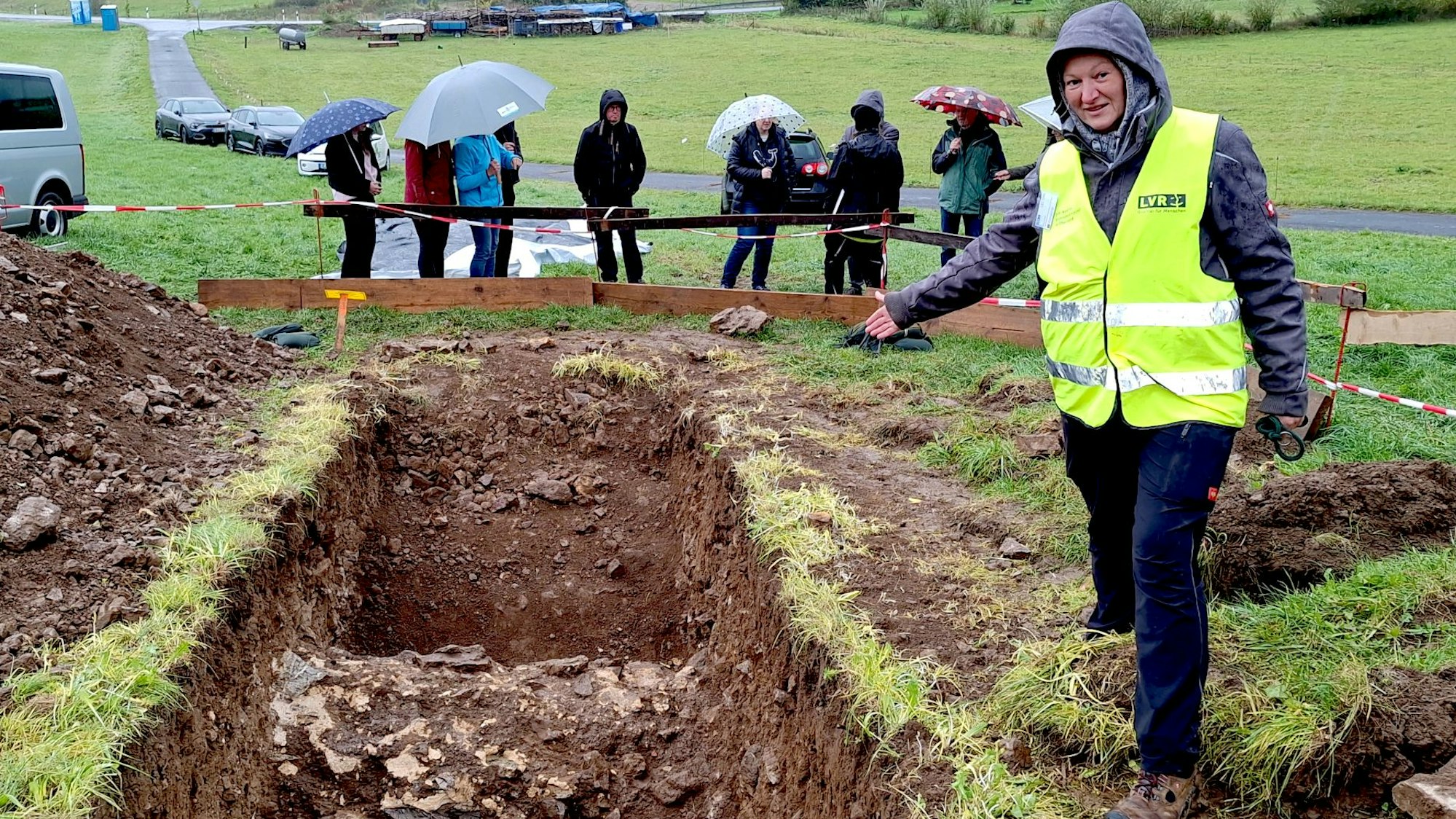 Eine Frau, die eine gelbe Warnweste über ihrer Kleidung trägt, deutet auf eine offene Ausgrabungsstelle auf einer Wiese, um die sich mehrere Besucher mit Regenschirmen versammelt haben.
